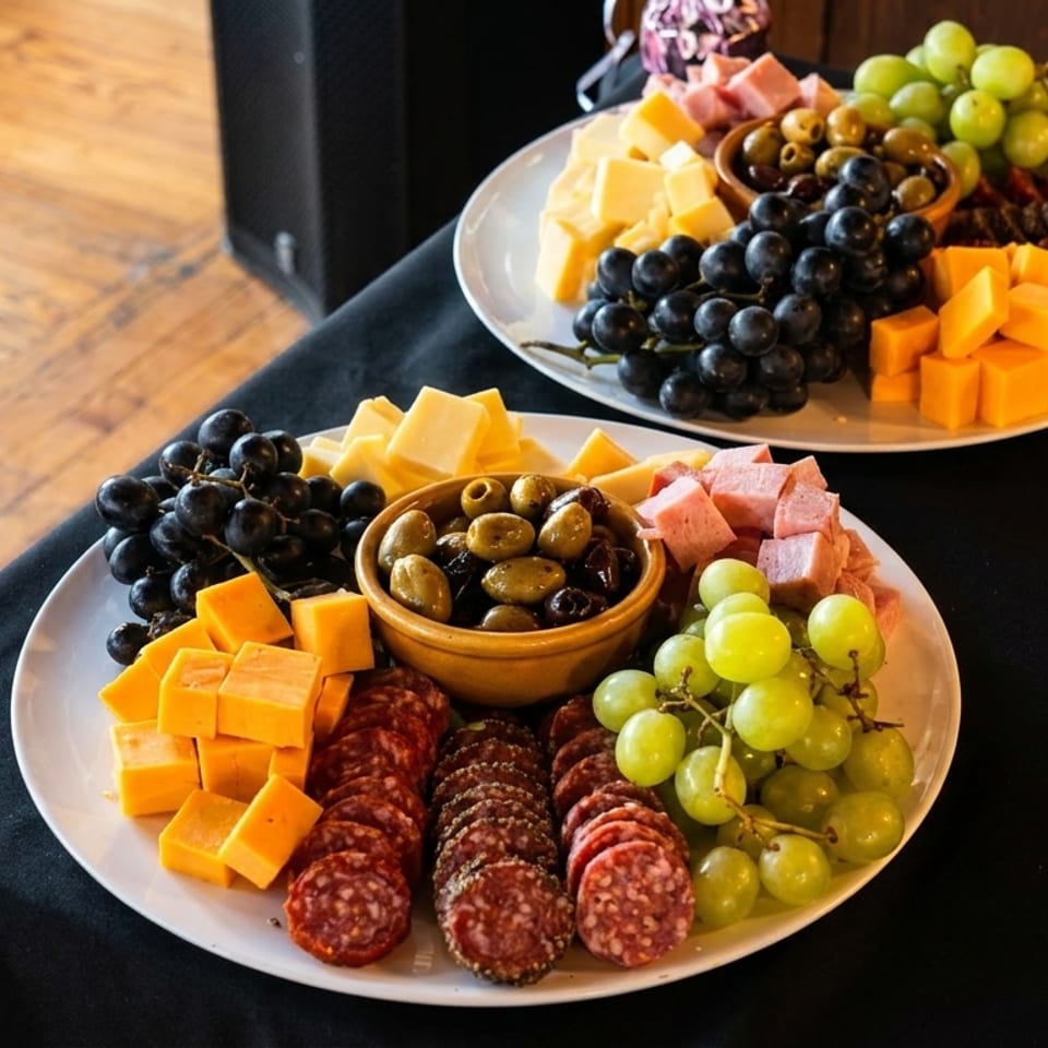 Charcuterie platters display assorted cheeses, sliced cured meats, grapes, olives, berries, and crackers on a black-draped table.