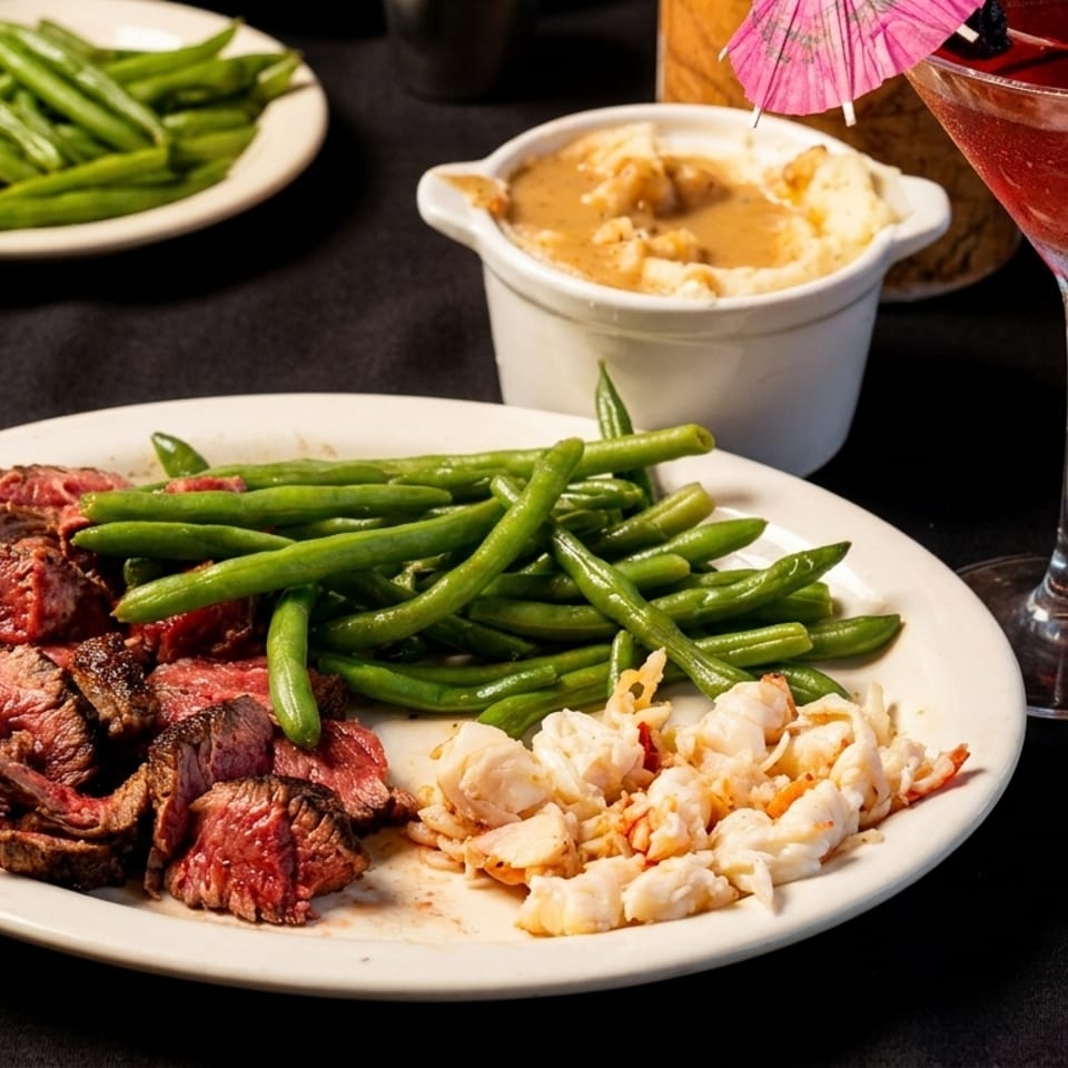 Plate of sliced medium-rare steak, green beans, and crab meat with mashed potatoes, alongside a red cocktail and pink umbrella.